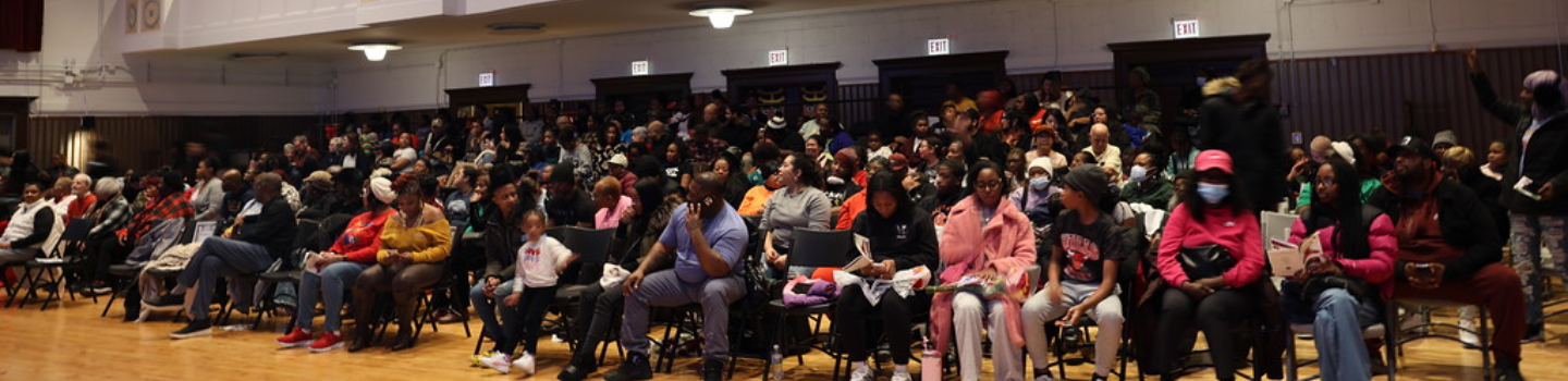 A large audience sits in folding chairs in an auditorium.